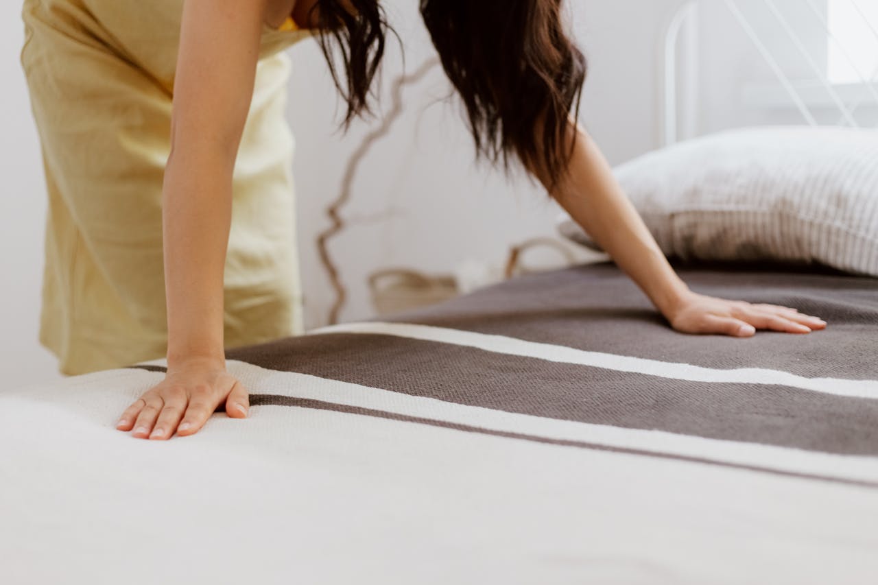 hero-img A woman making a bed adorned with striped linens in a bright, minimalist bedroom.